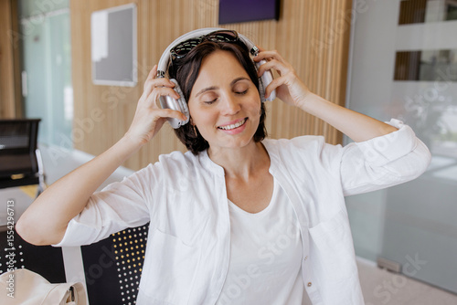 Happy woman sitting on a bench in a modern waiting area, smiling and listening to music with wireless headphones, relaxed traveler enjoying time in a contemporary public space, bus station