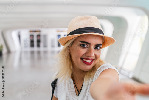 Smiling tourist wearing a stylish hat, reaching out with her hand at the bustling airport terminal, radiating excitement and cheerfulness
