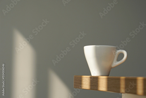 White coffee cup on wooden table under morning window light on clean grey wall background.