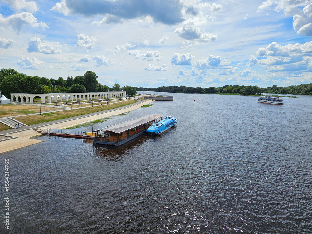 Naklejka premium Blue boat is docked next to a brown landing stage peer. The water is calm. There are several other boats in the water river Volkhov in Veliky Novgorod