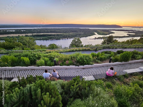 Group of people are sitting on a path overlooking a river. The sun is setting in the river Volga near Samara
