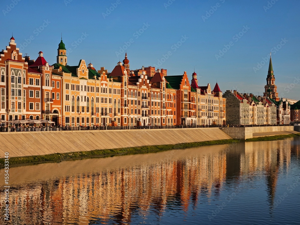 Naklejka premium City with a river running through it. The buildings are tall and colorful. The water is calm and reflects the buildings in Yoshkar-Ola - capital of the Republic of Mari El in Russia