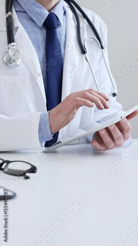 Male doctor wearing lab coat and stethoscope using digital tablet while sitting at desk in medical office, with medical chart and pen in the foreground