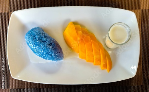 A top-down, close-up shot presents a classic Thai dessert, mango sticky rice, on a rectangular white plate. The dish features vibrant blue sticky rice, naturally colored with butterfly pea flower