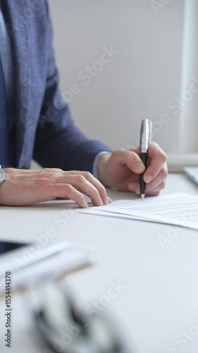 Close up of businessman's hands signing a contract with elegant pen, wearing a blue suit and a wristwatch, sitting at a white desk. Business people concept
