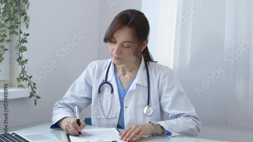 Female doctor wearing a lab coat and stethoscope, diligently writing notes on a clipboard while sitting at a desk in bright medical office with window and decorative plants. Medicine concept