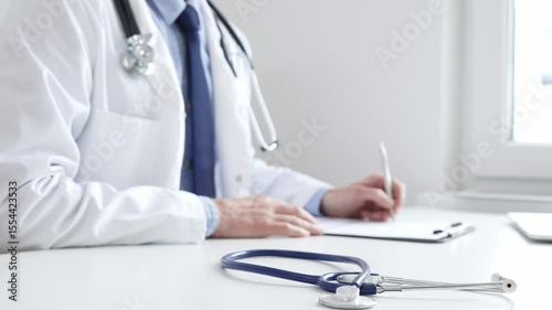Closeup of blue stethoscope is lying on the table near doctor man professional writing on the clipboard in clinic office. Medicine and healthcare