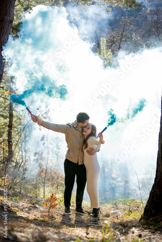 Cheerful joyfully celebrating their pregnancy couple surrounded by blue smoke in a beautiful, lush forest
