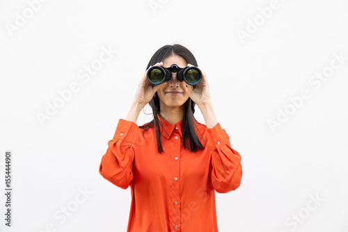 Woman in orange shirt smiling and using binoculars against a white wall