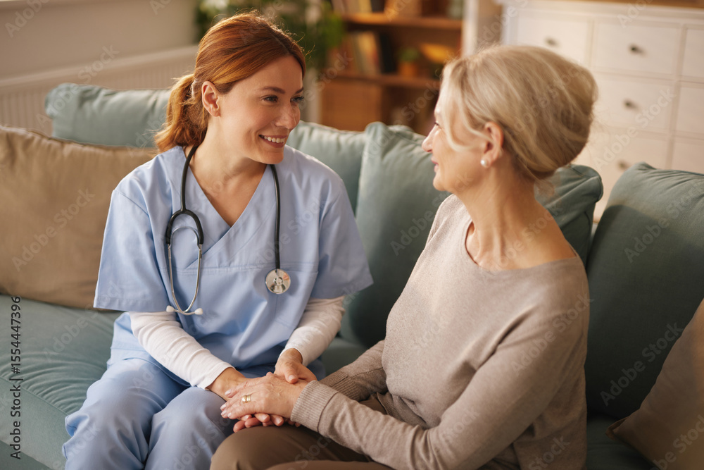Fototapeta premium A smiling nurse in scrubs and a stethoscope provides care and support to a senior woman in her living room. They are sitting on a couch together, holding hands, and looking at each other.