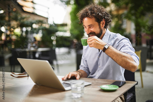 Obraz na plátně Man engaged in work on his laptop, smiling while sipping coffee at an outdoor cafe, surrounded by greenery and other patrons enjoying the pleasant weather