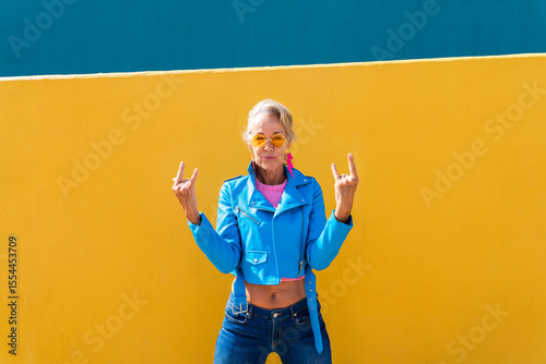 Mature woman gesturing horn sign in front of colored wall