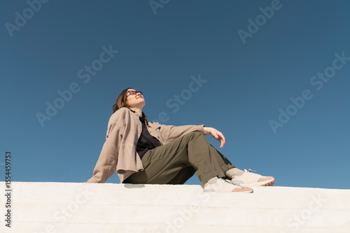 Woman sitting on steps outdoors under a clear blue sky, looking relaxed and fashionable