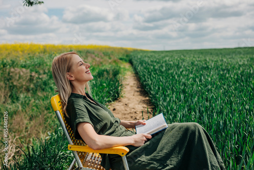 Woman sitting on a yellow folding chair in a field, reading a book and enjoying the countryside.