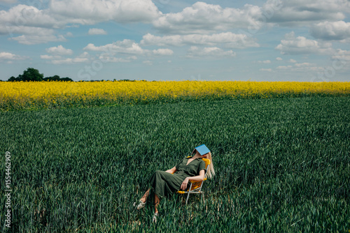 Woman relaxing on a yellow folding chair in a field of green wheat and yellow rapeseed, reading a book under a cloudy sky