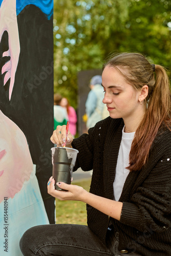 Young brunette woman painting large canvas outdoors. Focused and serene, embodying artistic expression. Green foliage creates vibrant backdrop