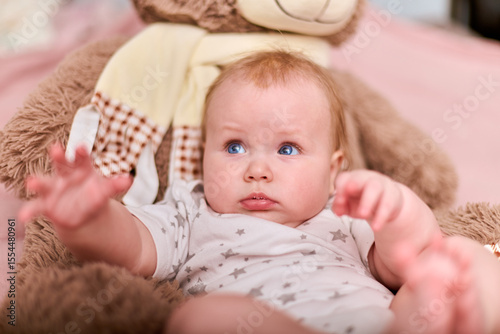 Young female with light brown hair and blue eyes on pink blanket. Wearing white star-patterned outfit, reaching towards camera on teddy bear chair, curious emotion