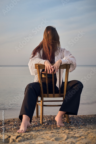 Young woman sits on chair facing calm sea, symbolizing solitude and introspection. Her relaxed posture and flowing hair evoke serene, contemplative mood at dusk