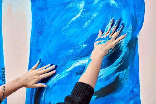 Young female with fair hair, engaging in expressive painting using hands. Dynamic blue strokes cover large surface, with vibrant energy and creativity in focus