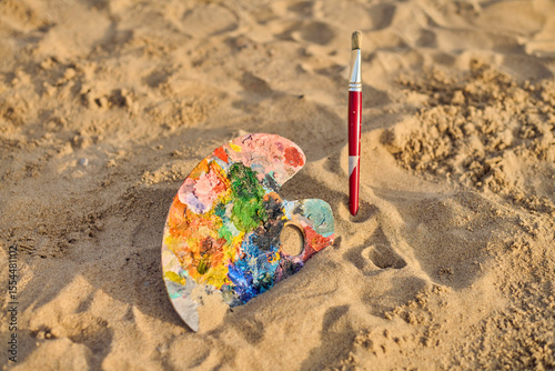 Vibrant artist palette and paintbrush lie on sandy beach, blending creativity with nature. Setting evokes sense of freedom and inspiration, under warm sun