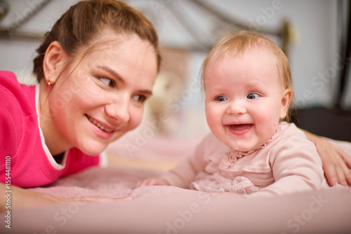 Young mother with brown hair, smiling baby girl indoors. Warm pink tones, soft lighting, metal bedframe in background. Expression of joy and connection