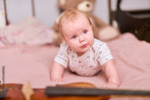 Baby girl with blonde hair lies on pink bed, curiously reaching for violin. Cozy bedroom setting, warm lighting enhances peaceful and playful atmosphere