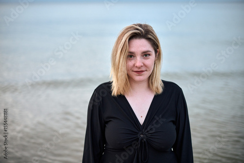 Young woman with blonde hair and serene expression stands by sea. Overcast sky and gentle waves create tranquil and introspective mood