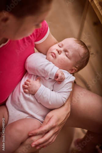 Young woman with dark hair in pink shirt holds sleeping baby girl. Infant in white clothing rests peacefully, enveloped by warm indoor lighting. Soft background create cozy atmosphere