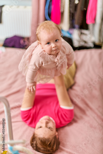 Young mother with brown hair and fair skin plays with baby girl in pink dress on bed. Room has colorful clothes in background. Joyful and cozy scene