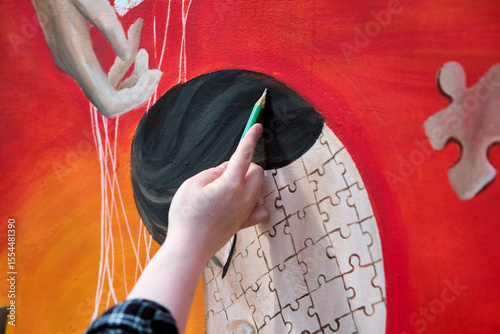 Close-up of adult female hand painting puzzle-themed mural. Vibrant red dominates, with canvas of puzzle pieces. Daylight highlights intricate detail