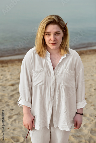 Young woman stands on serene beach, paintbrush in her hair, evoking creativity and tranquility. Her white shirt contrasts with calm sea, capturing contemplative elegance under soft daylight