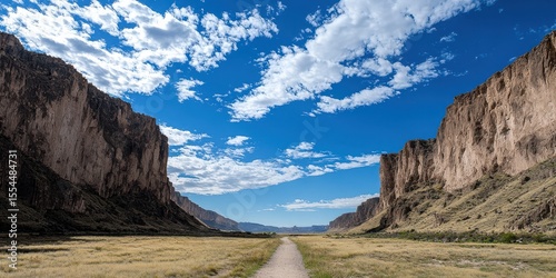 A scenic canyon with towering cliffs on both sides, a dirt path in the middle, and a vibrant blue sky filled with scattered clouds.