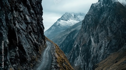 Narrow mountain path winding between steep rocky cliffs with a snow-capped peak in the background under an overcast sky.