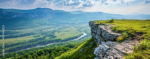 Scenic view of a rocky cliff overlooking a winding river, lush green forests, and distant mountains under a partly cloudy sky.