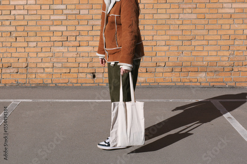 Young woman holding blank white tote bag canvas fabric, city street, brick wall background. Female consumer hold big white textile shopper. template mock up design for text or logo place