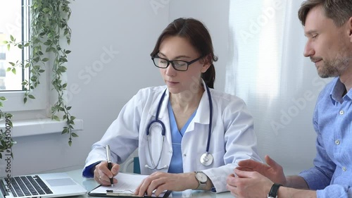 Female doctor taking notes on clipboard while listening attentively to patient during medical consultation in hospital office, promoting communication and understanding.