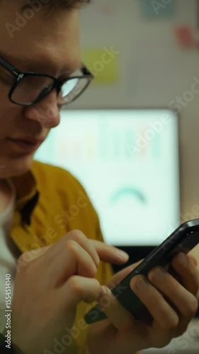 A young man in glasses engages with his smartphone in a cluttered home office at night. The atmosphere is focused and productive, with a computer screen displaying data in the background