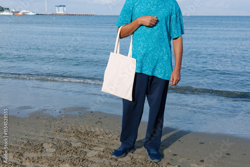 Man with black, tan skin holding in hand white eco cotton textile bag. Blue water of sea or ocean coast background. Seaside, sunny day. Blank mock up tote bag, eco concept, place for text design logo