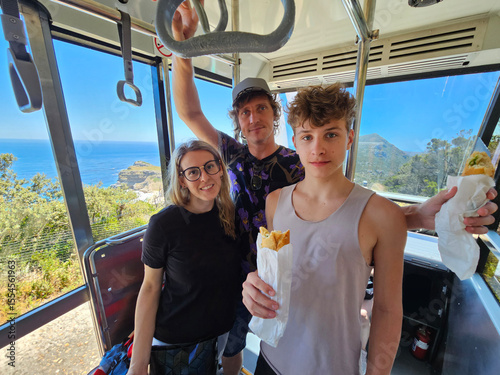 Man in a black shirt with the word super on it is holding a bag. A woman and a boy are standing next to him in cabin in Cape of good hope in South Africa