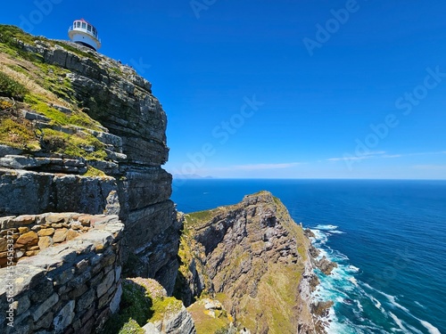 Lighthouse is on top of a cliff overlooking the ocean. The sky is clear and blue. The ocean is calm and the water is blue in Cape of good hope in South Africa