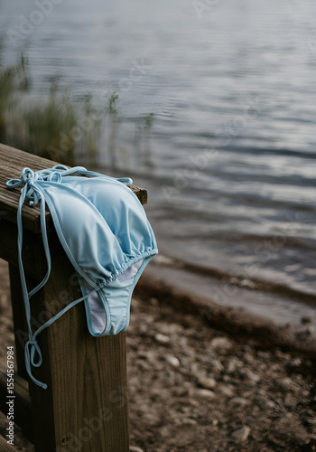 A light blue bikini casually draped over a rustic wooden deck railing, captured beside a calm lake shoreline with visible pebbles and sparse vegetation.