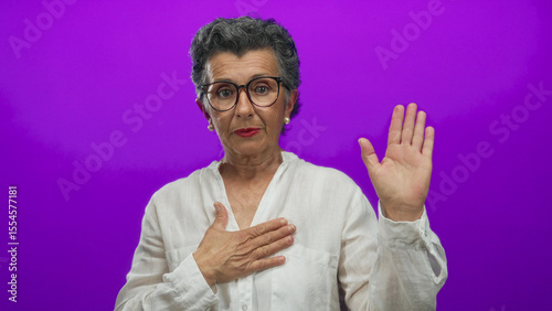 Senior woman with grey hair making a gesture of oath against a vibrant purple background, wearing glasses and a white shirt, conveying sincerity and commitment.
