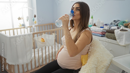 Canvastavla Pregnant woman sitting in a baby room drinking water with a cradle behind her, conveying a serene mood and highlighting indoor motherhood moments in a cozy home environment