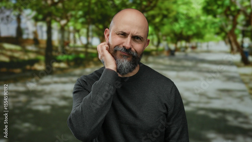 Man with beard standing in park outdoor on sunny day surrounded by green trees exuding a contemplative mood with hands partially over face and wearing casual dark attire