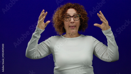 Woman gesturing humorously with expressive face against blue background wearing glasses and white shirt conveying fun emotion in isolated setting