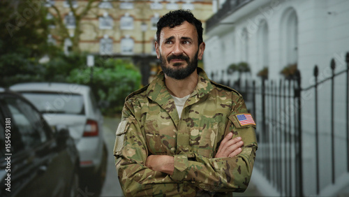 Man with beard wearing military uniform stands outdoors in city street with cars, showcasing a serious expression.