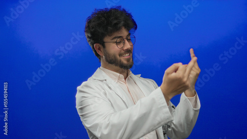Young man with a beard wearing a white coat humorously gestures with middle fingers against a solid blue background, smiling and exuding a playful yet rebellious attitude.