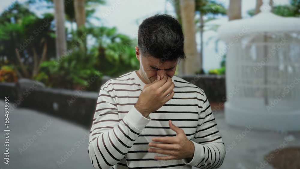Fototapeta premium Young man outdoors in a city setting with a pained expression pinching his nose surrounded by lush green foliage and a white gazebo in the background.