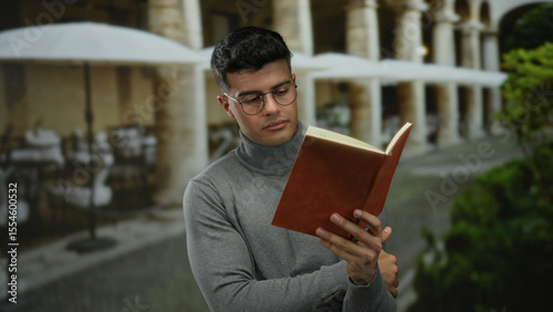 Young man in glasses reads a book on a terrace at an outdoor city restaurant, surrounded by greenery and umbrellas, providing a serene and studious ambiance.