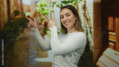 Free Download Woman smiling in white sweater pointing with fingers on outdoor city street with green foliage and stone walls. - GFXLoad